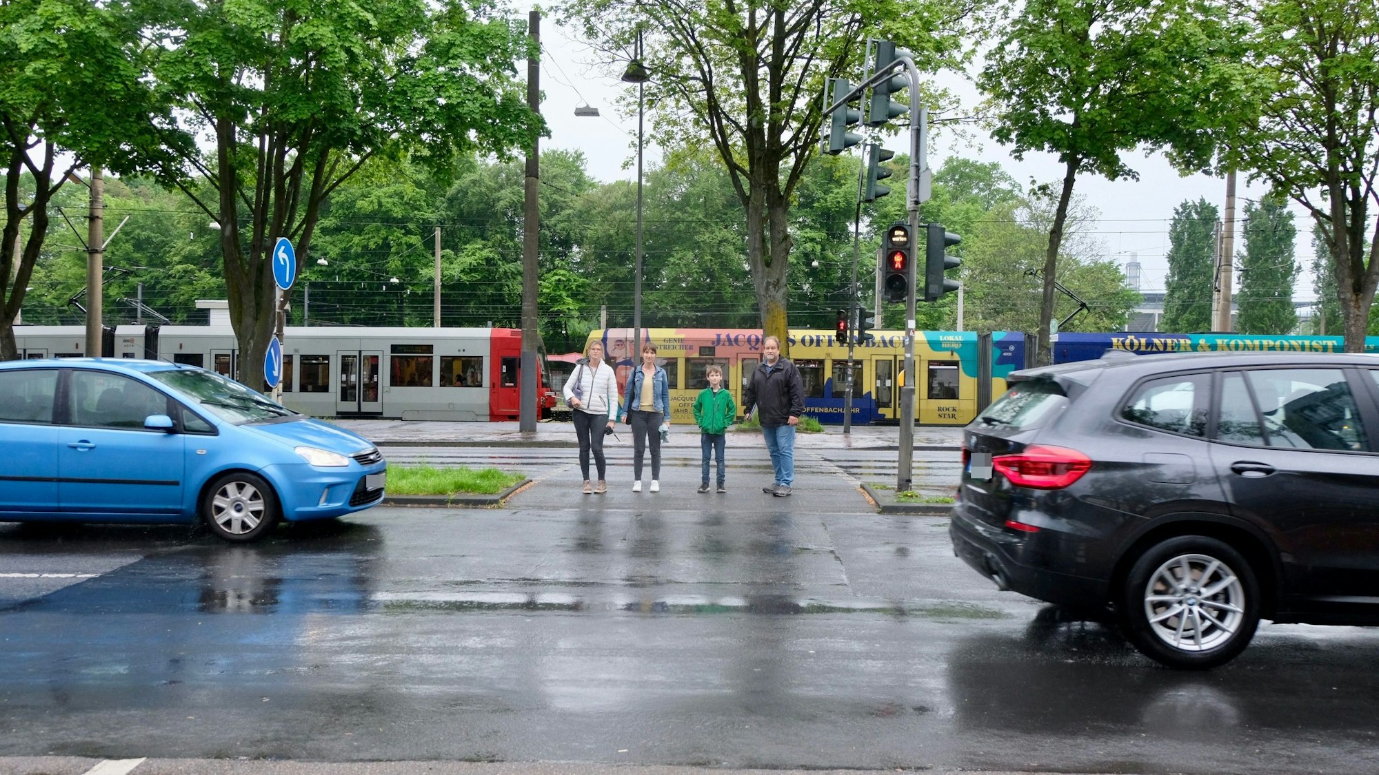 Vier Personen stehen an einer Kreuzung, auf der Straße fahren Autos.