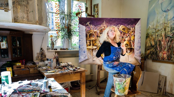 Eine Künstlerin posiert in einem Atellier vor einem bunten Bild auf einer Leinwand.