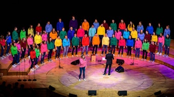 Der Jugendchor St. Stephan beim Auftritt in der Kölner Philharmonie. Kinder und Jugendliche stehen in drei Reihen aufgereit vor dem Chorleiter, alle tragen bunte T-Shirts in unterschiedlichen Farben.