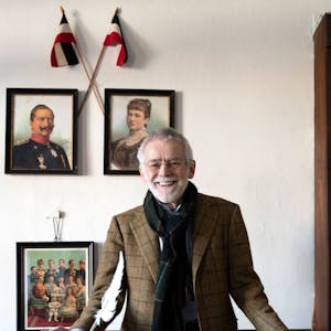Dr. Peter Joerißen, Leiter Schulmuseum Bergisch Gladbach, im alten Klassenzimmer der ehemaligen Volksschule. Er steht vor einigen Gemälden, die an einer weißen Wand hängen, und lächelt an der Kamera vorbei.