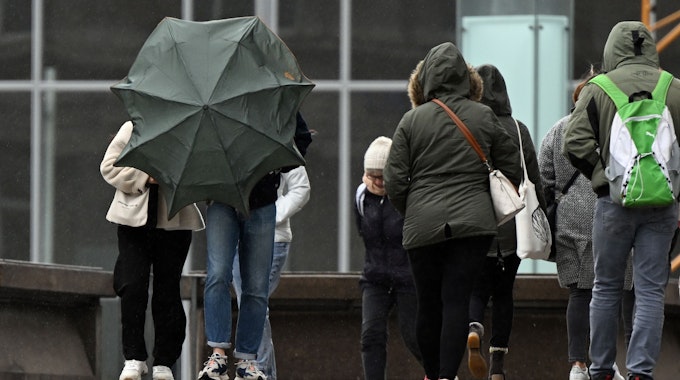 Spaziergänger kämpfen auf der Domplatte mit ihrem Regenschirm (Archivbild vom April 2022): Nach einem freundlichen Vormittag soll es am Sonntagnachmittag und -abend in NRW laut Vorhersage ungemütlich werden.