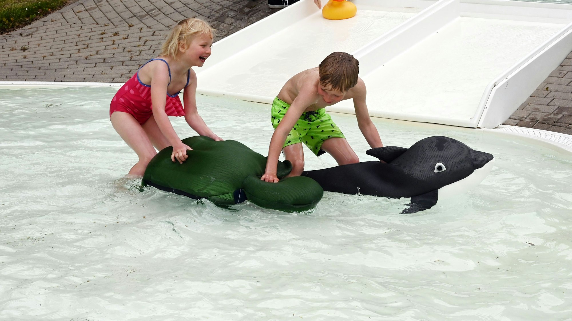 Im Bild sind ein Junge und seine kleine Schwester, die in einem Schwimmbecken für Kinder mit zwei aufblasbaren Tieren spielen.