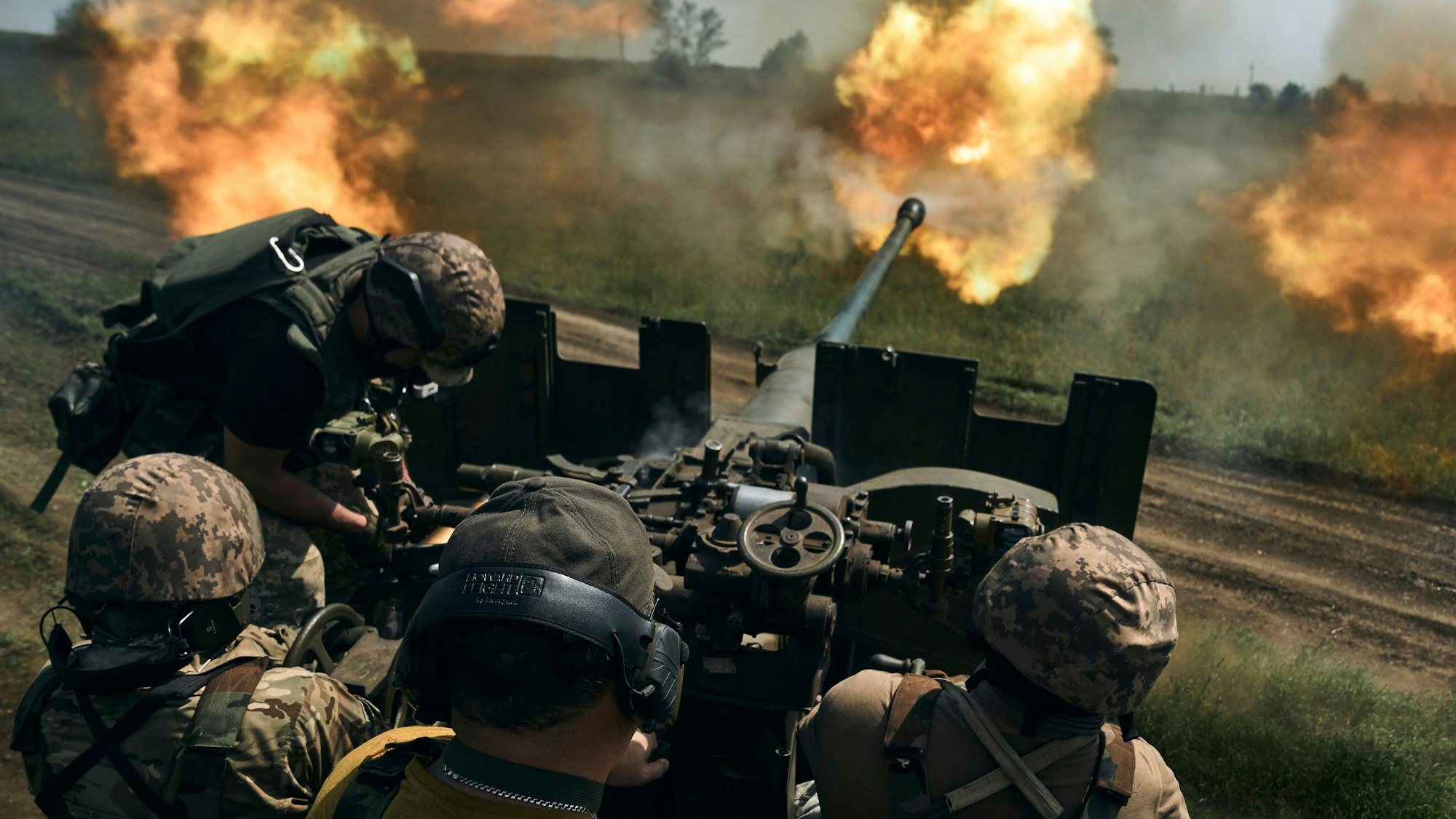 Männer in Tarnuniformen sitzen hinter dem Zielrohr einer Kanone, aus deren Mündung Feuer kommt.