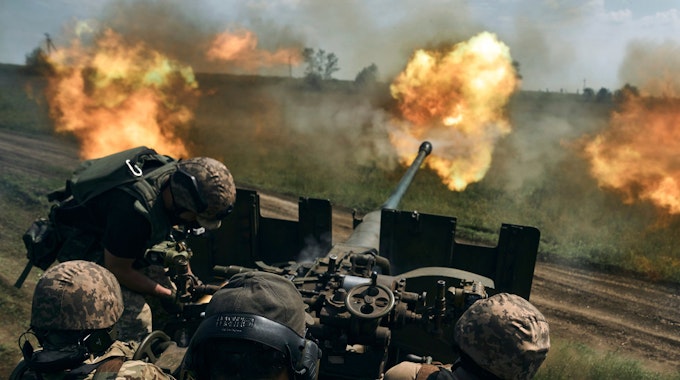 Männer in Tarnuniformen sitzen hinter dem Zielrohr einer Kanone, aus deren Mündung Feuer kommt.