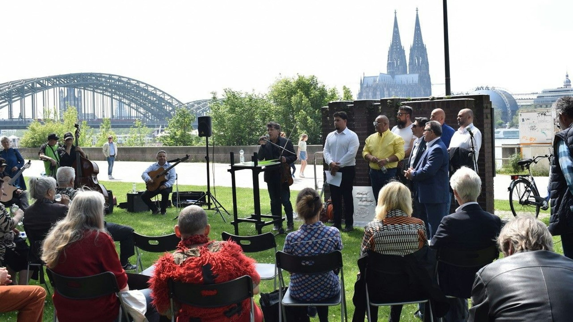 Markus Reinhardt spricht vor mehreren Zuhörern am Deutzer Rheinufer, im Hintergrund sind der Rhein und Dom zu sehen.