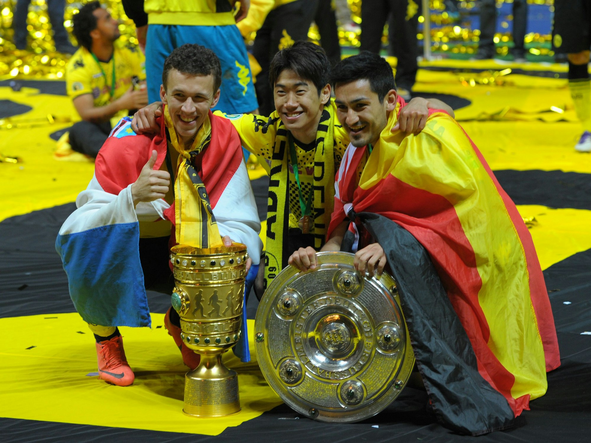 Ivan Perisic, Shinji Kagawa und Ilkay Gündogan posieren hockend mit dem Pokal und der Meisterschale.Perisic ist in einem Kroatien-Flagge gehüllt, Gündogan in eine Deutschland-Fahne.