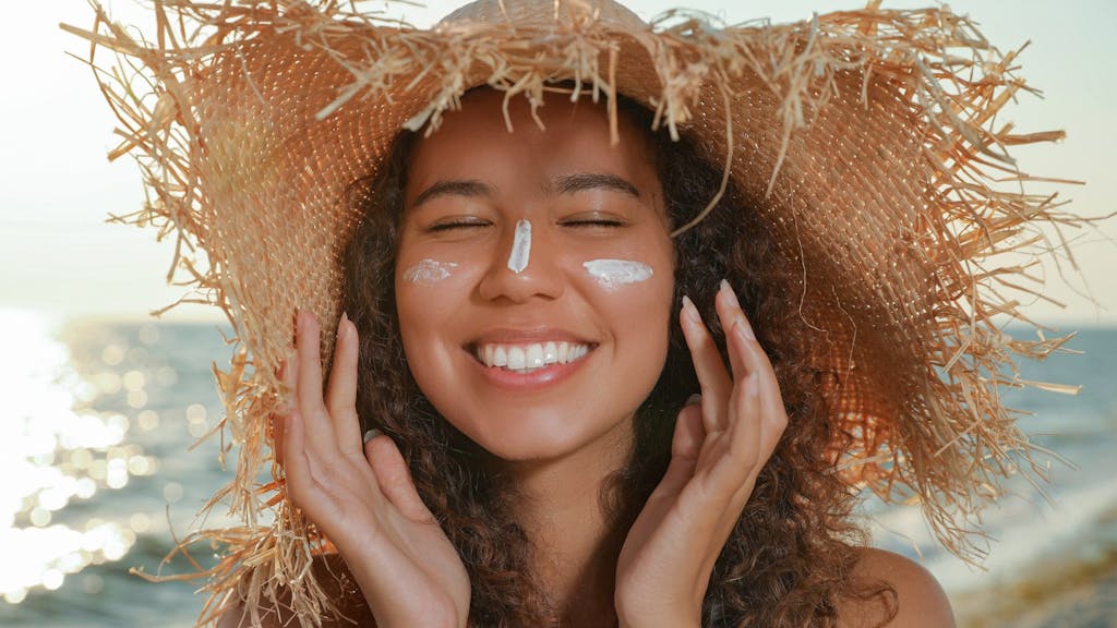 Lächelnde Frau mit Sonnensutzcreme im Gesicht und Strandhut auf dem Kopf. 