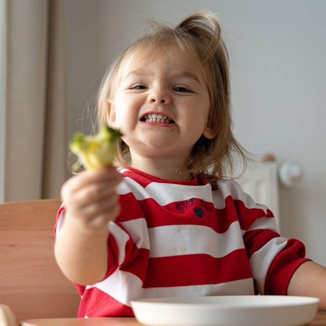 Ein Kleinkind sitzt in einem Hochstuhl und isst Brokkoli.