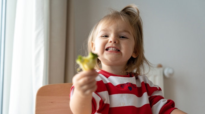 Ein Kleinkind sitzt in einem Hochstuhl und isst Brokkoli.
