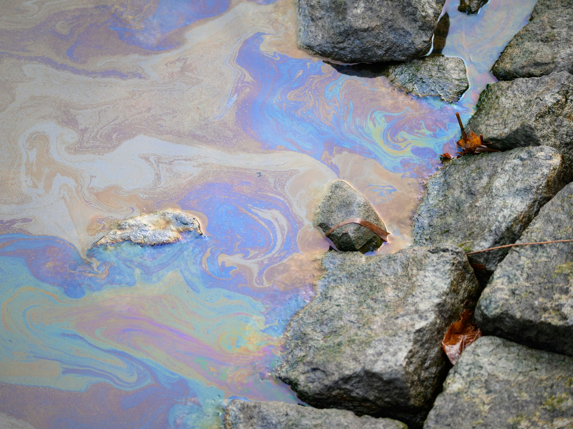 Ein Ölfilm in Regenbogenfarben, daneben liegen Steine.