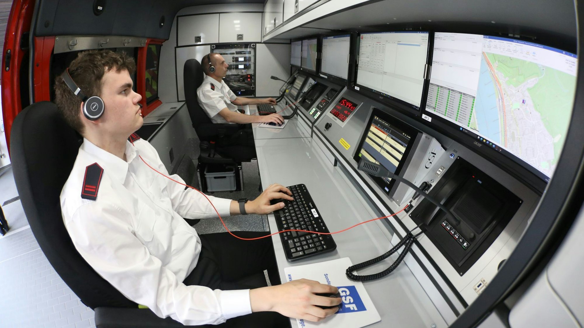 Zwei Männer sitzen vor Computermonitoren im Einsatzleitwagen der Feuerwehr und bedienen Maus und Tastatur. Beide tragen Headsets.