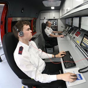 Zwei Männer sitzen vor Computermonitoren im Einsatzleitwagen der Feuerwehr und bedienen Maus und Tastatur. Beide tragen Headsets.&nbsp;
