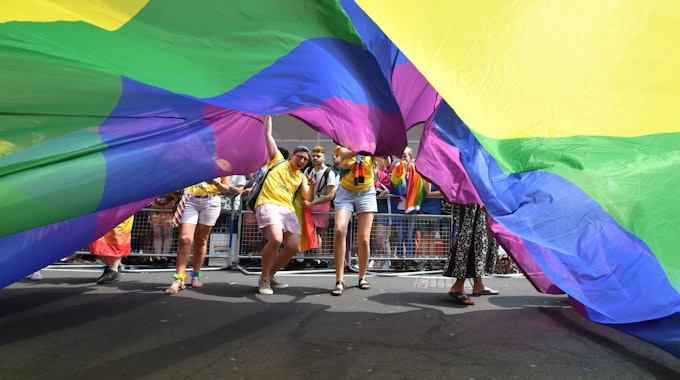 Teilnehmende der Londoner Pride Parade 2018 schauen unter einer riesigen Regenbogenflagge hervor.
