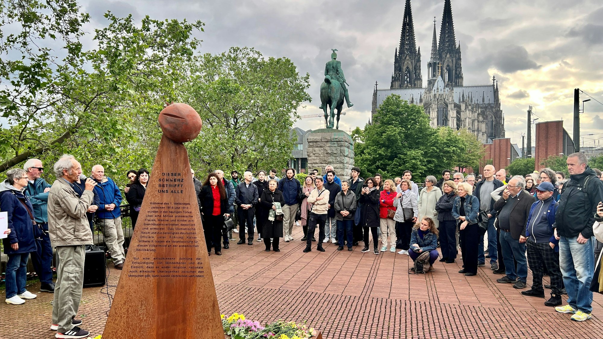 Menschen am Armenier-Mahnmal an der Hohenzollernbrücke demonstrieren gegen den geplanten Abbau der Skulptur im Bildvordergrund. Im Hintergrund ist der Kölner Dom zu sehen.