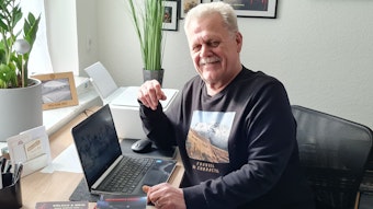 Ein älterer Mann mit Schnurrbart sitzt im Pullover vor einem Laptop und grinst in die Kamera.