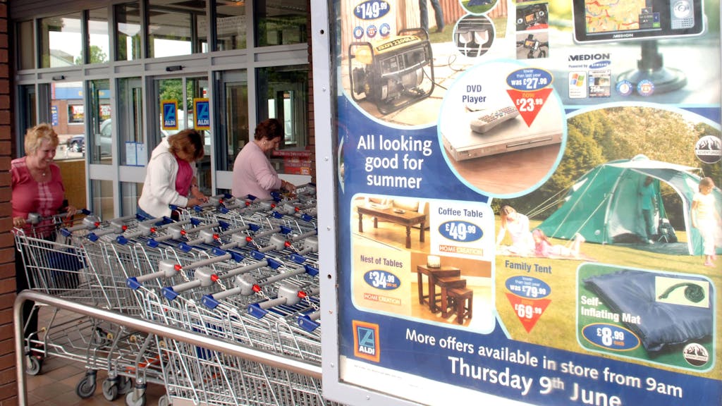 Aldi-Süd-Plakat mit Werbung an einer Filiale in England neben drei Frauen an Einkaufswagen