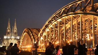Menschen stehen in der Nacht vor der beleuchteten Hohenzollernbrücke.