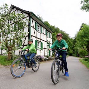 Unten am Mühlenweg ist die Radwelt noch in Ordnung, aber von hier führen nur steile Wanderwege hinauf nach Glöbusch. Zwei Kinder sitzen auf Fahrrädern.