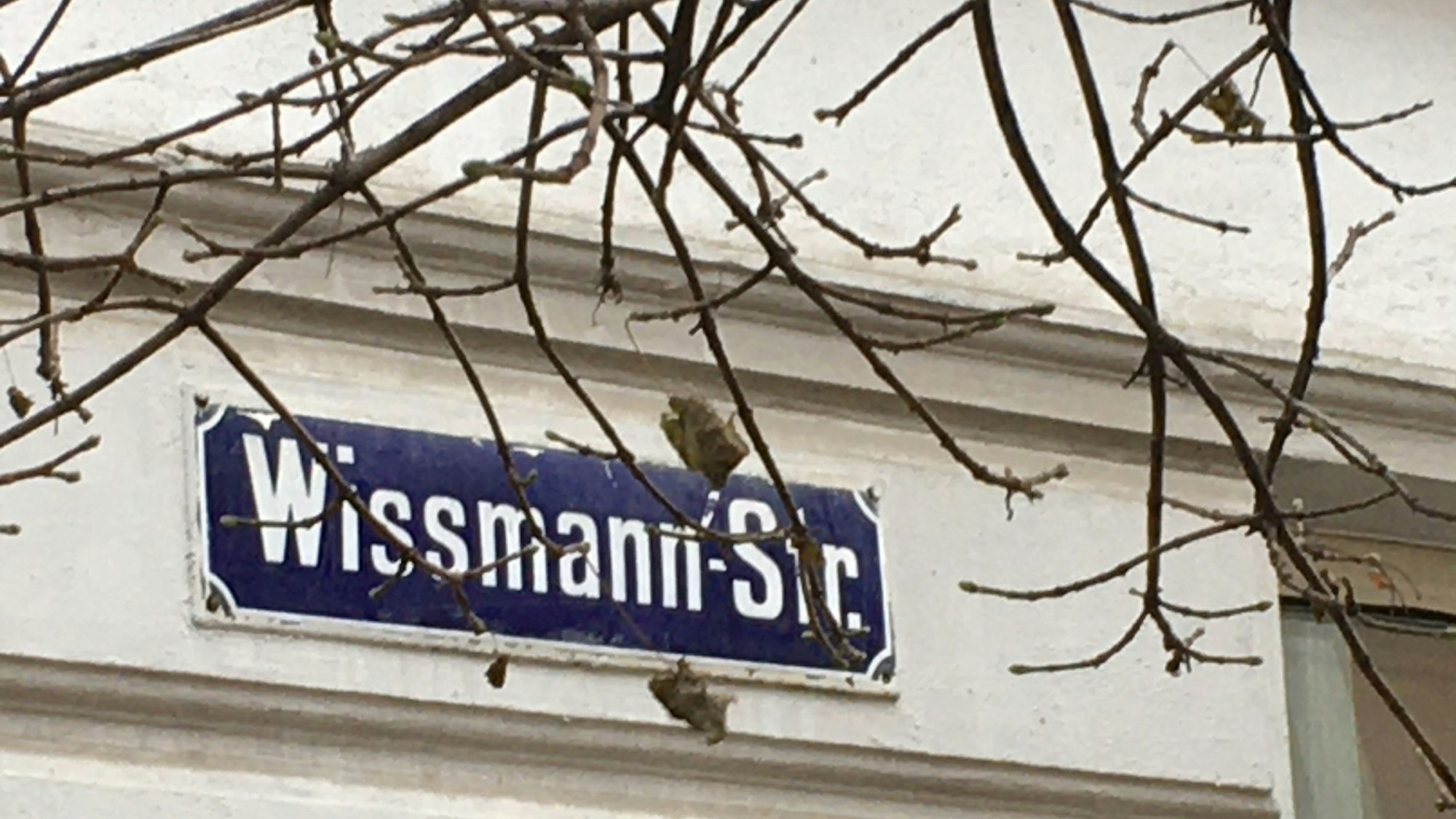 Ein Straßenname auf einem blauen Schild hängt an einer Hauswand.