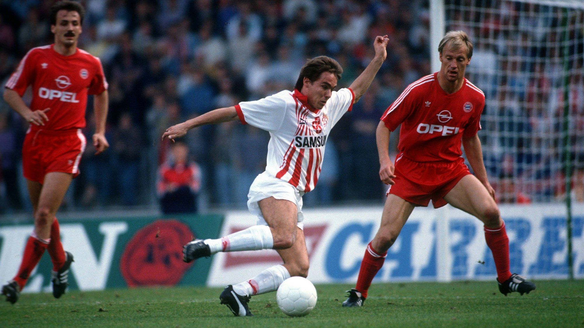 Der Kölner Spieler Olaf Janßen im weißen Trikot im Duell mit einem in rot gekleideten Gegenspieler des FC Bayern München.