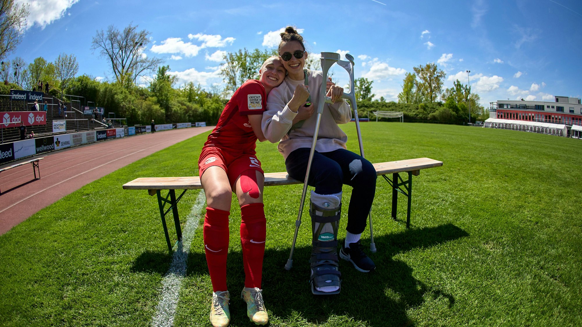 Fußballerin Gianna Rackow sitzt mit Krücken sitzt gemeinsam mit einer Mitspielerin auf einer Bank in einem Stadion.