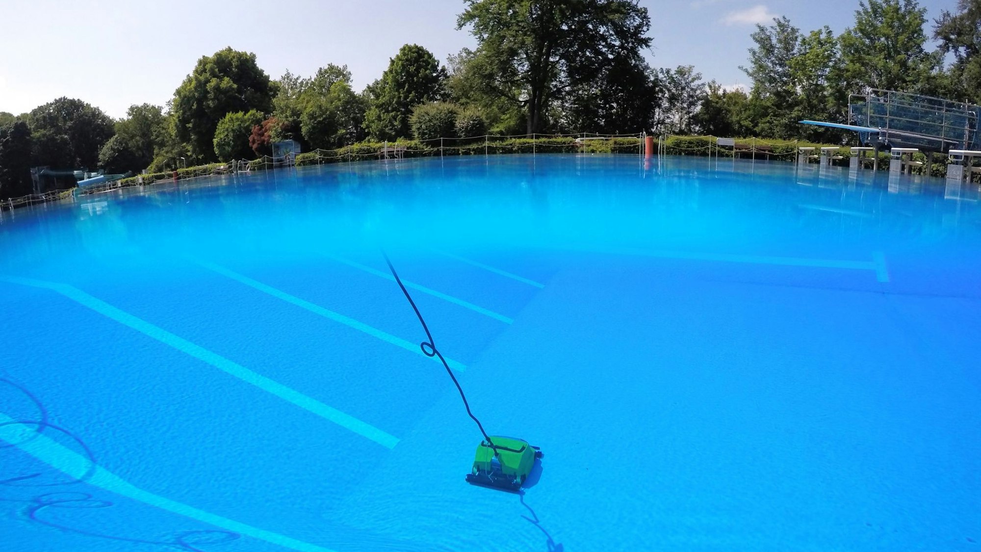 Im blauen Wasser zieht auf dem Boden des 50-Meter-Beckens eine gelbe Reinigungsmaschine ihre Bahnen. Am oberen Bildrand sind Bäume und Sträucher zu sehen, rechts am Rand ein Sprungbrett.