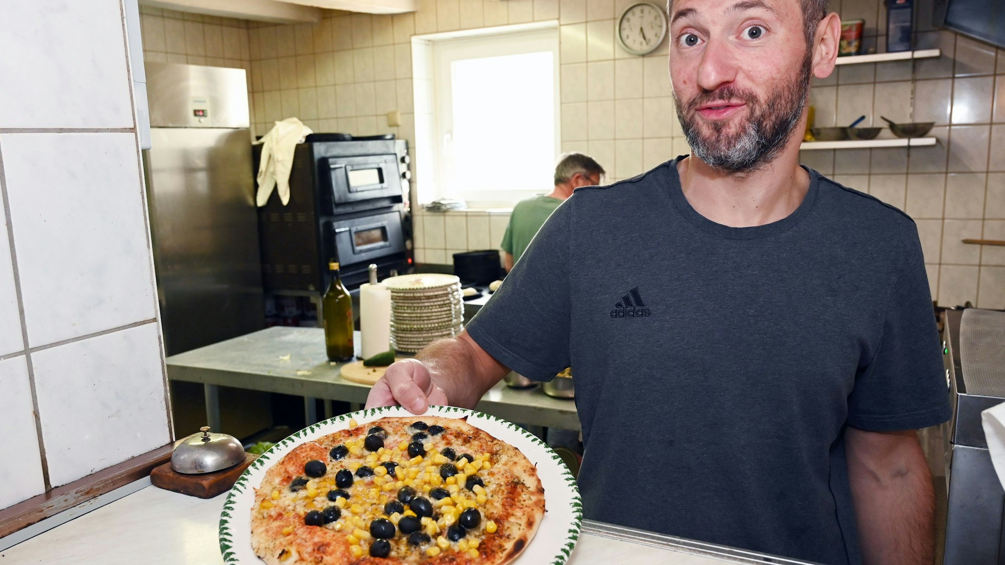Pizzabäcker und Bayern-Fan Luigi Zeni zeigt eine Pizza mit gelbem Mais und schwarzen Oliven.