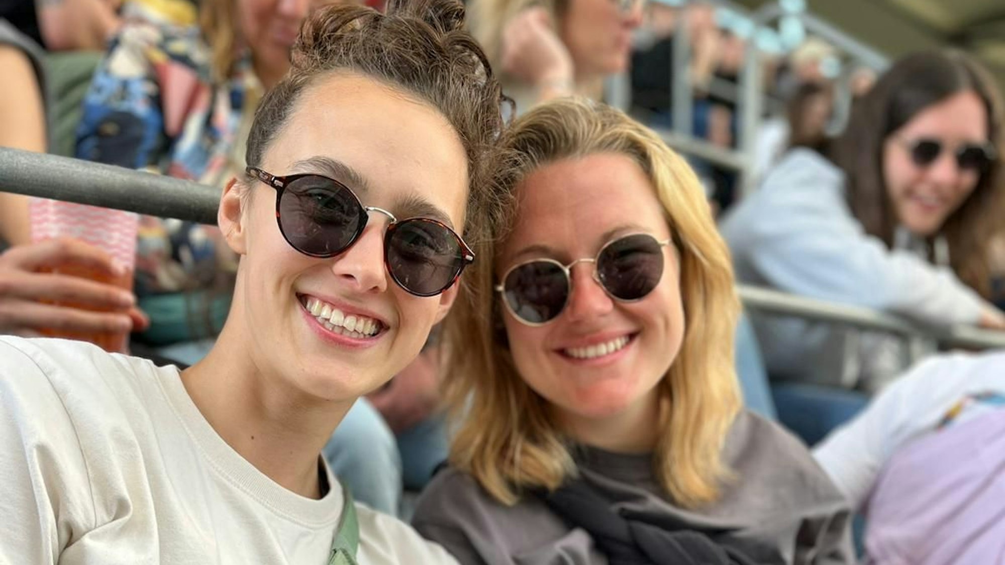 Zwei junge Frauen sitzen in einem Stadion nebeneinander und lachen in die Kamera.