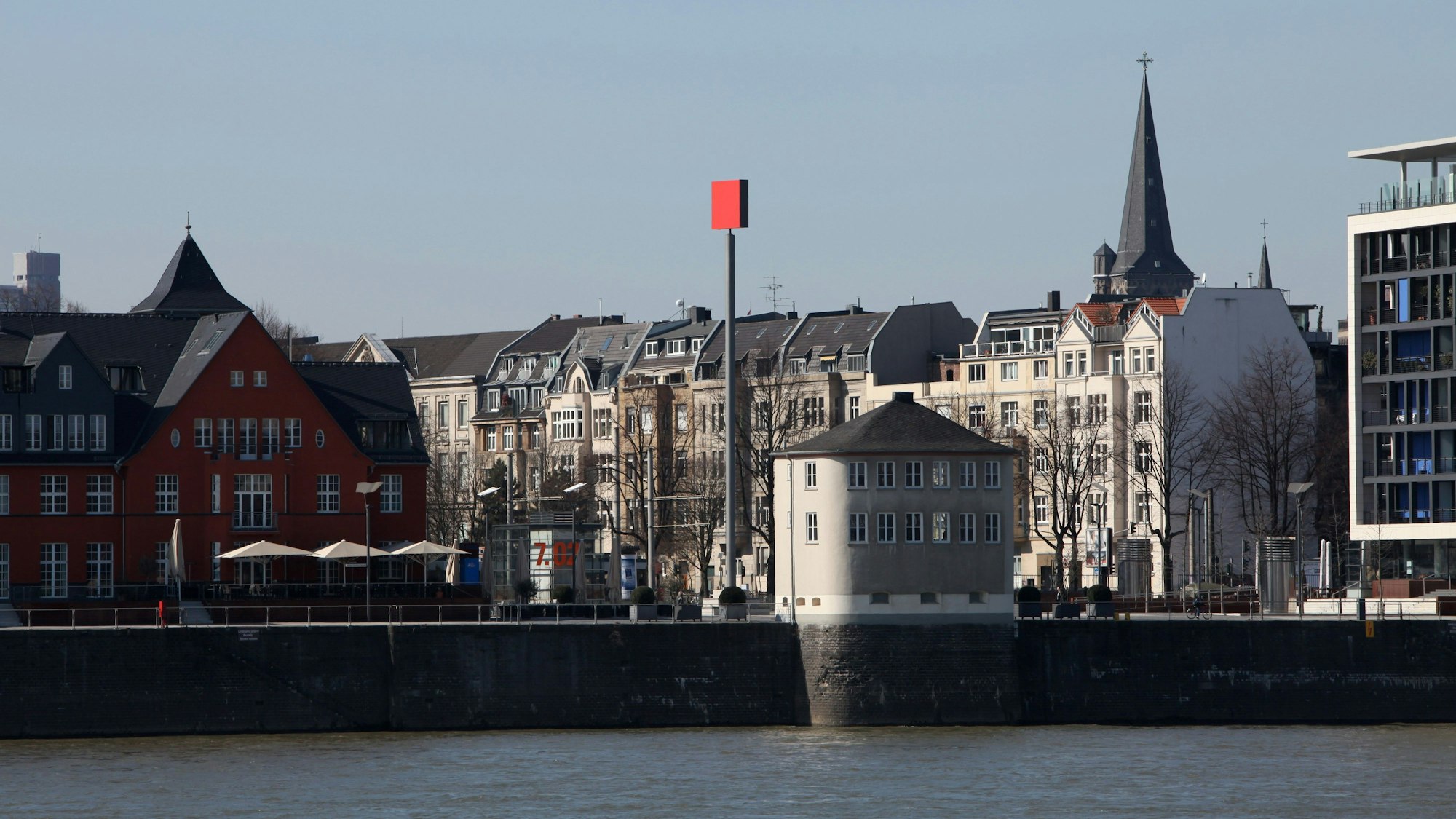 Ein rotes Quadrat auf einer Stele steht vor der Silhouette der Häuser am Rheinauhafen.