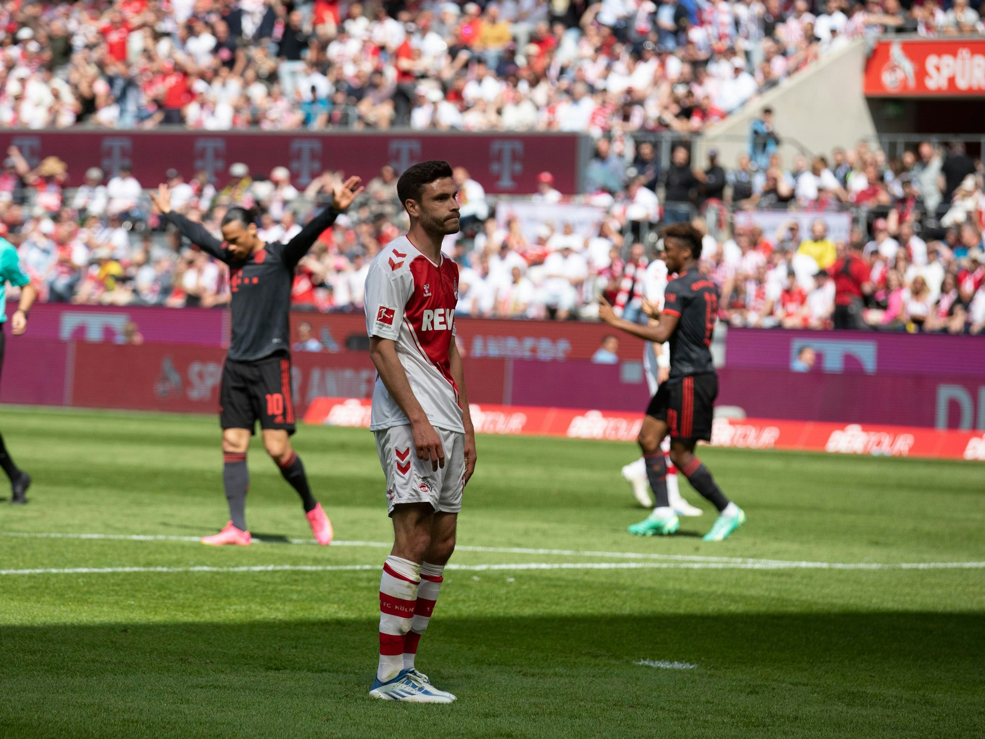 Jonas Hector steht auf dem Platz, seine Schultern hängen nach dem Gegentor durch die Bayern. Im Hintergrund jubeln Leroy Sané und Kingsley Coman.