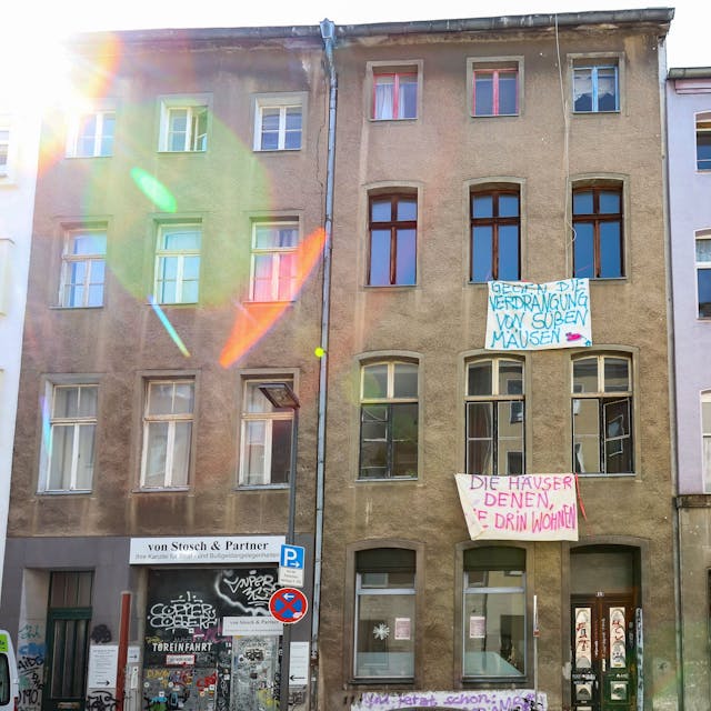 Eine Häuserzeile mit vier mehrgeschossigen Häusern ist zu sehen. Vor der Front des braunen Hauses hängen Banner mit Slogans, etwa Die Häuser denen, die drin wohnen.&nbsp;