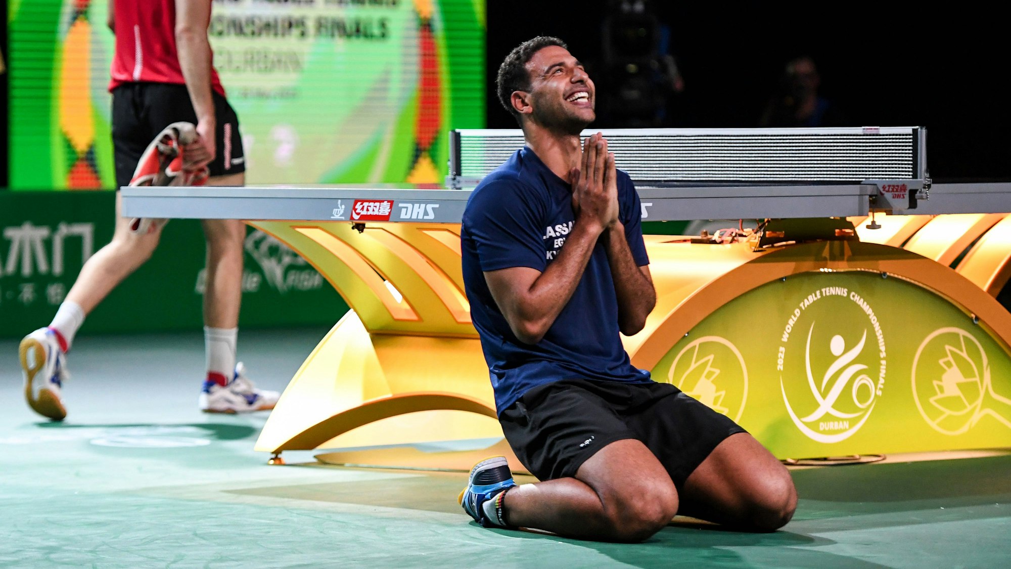 Ein Tischtennisspieler kniet mit betenden Händen auf dem Boden vor einer Tischtennisplatte und schaut lachend nach oben.