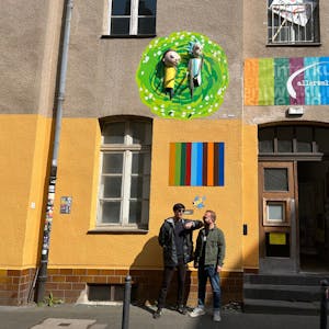 Yaşar Fidan (l.) und Lemi Karaca von Coach e.V. stehen vor ihrem Kunstwerk am Allerweltshaus in Ehrenfeld. Die 3D-Installation zeigt Rick und Morty aus der gleichnamigen Serie, wie sie aus einem Portal kommen.