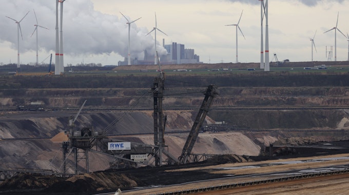 Der Blick auf den Braunkohletagebau Garzweiler II. zeigt schwere Maschinen der RWE im Fordergrund, die Rauch ausstoßen und Windräder im Hintergrund vor einem bewölkten Himmel.