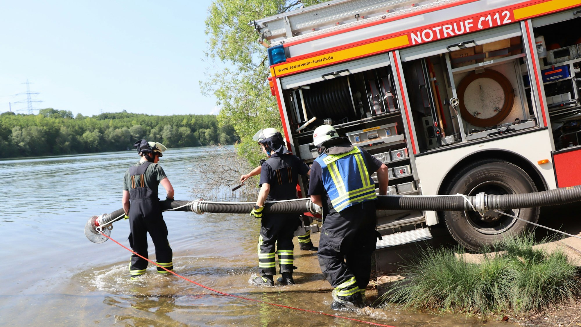 Ein Feuerwehrauto steht am Rand des Sees, Feuerwehrleute verlegen einen Schlauch ins Wasser.