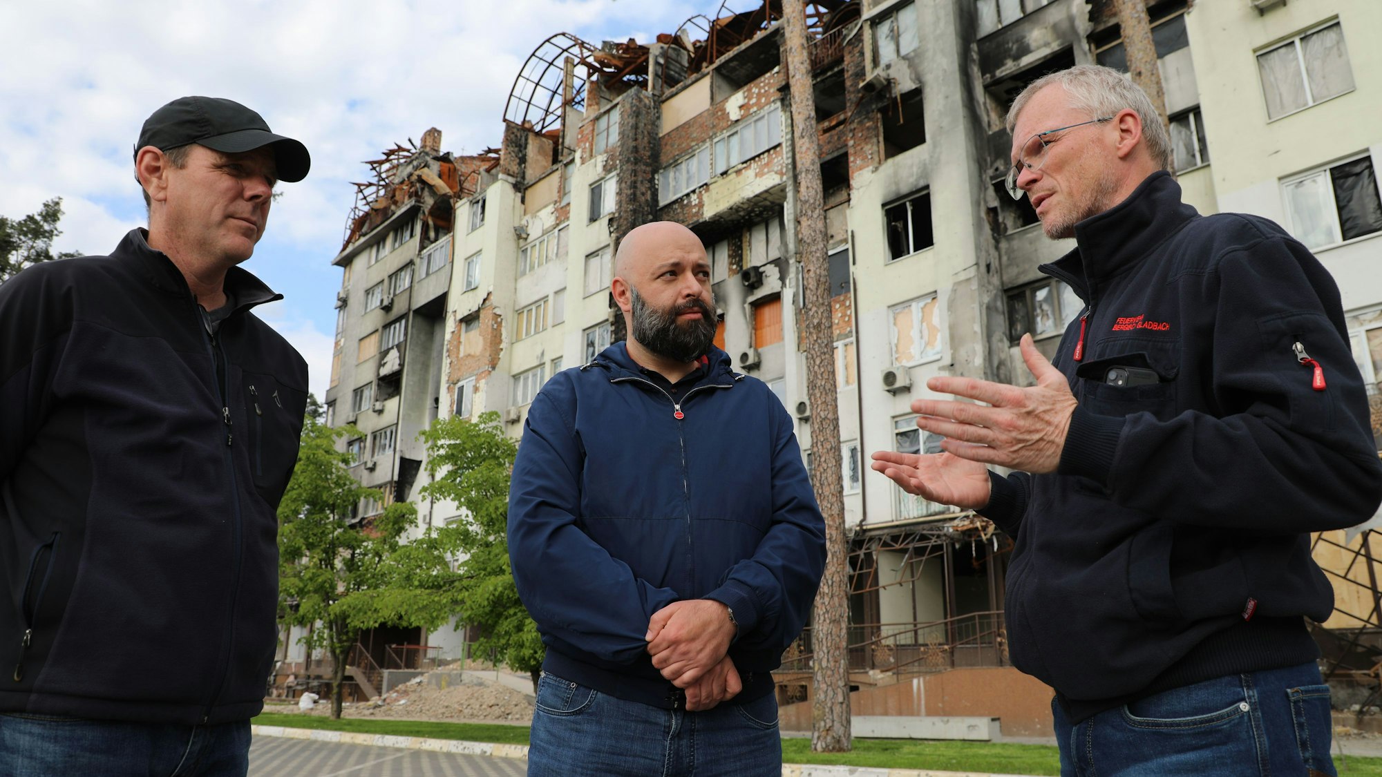 Drei MEnschen stehen sprechend vor einem zerbombten und ausgebrannten Wohn- und Geschäftshaus bei Butscha.