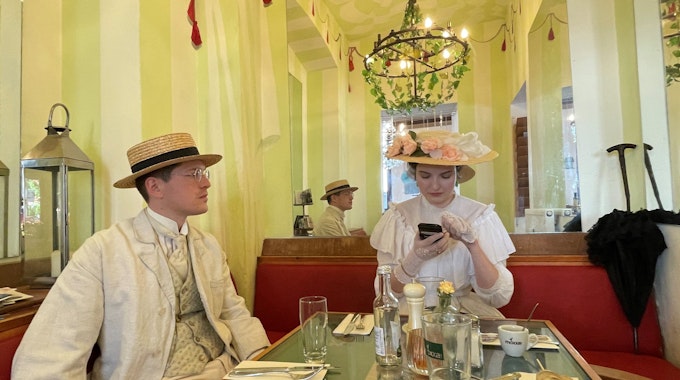 Ein Mann mit Hut und hellem Anzug mit Weste und Krawatte und eine Frau mit einem mit Blumen besetztem Hut auf dem Kopf und einem rüschenbesetzten weißen Kleid sitzen an einem Tisch in einem Café.