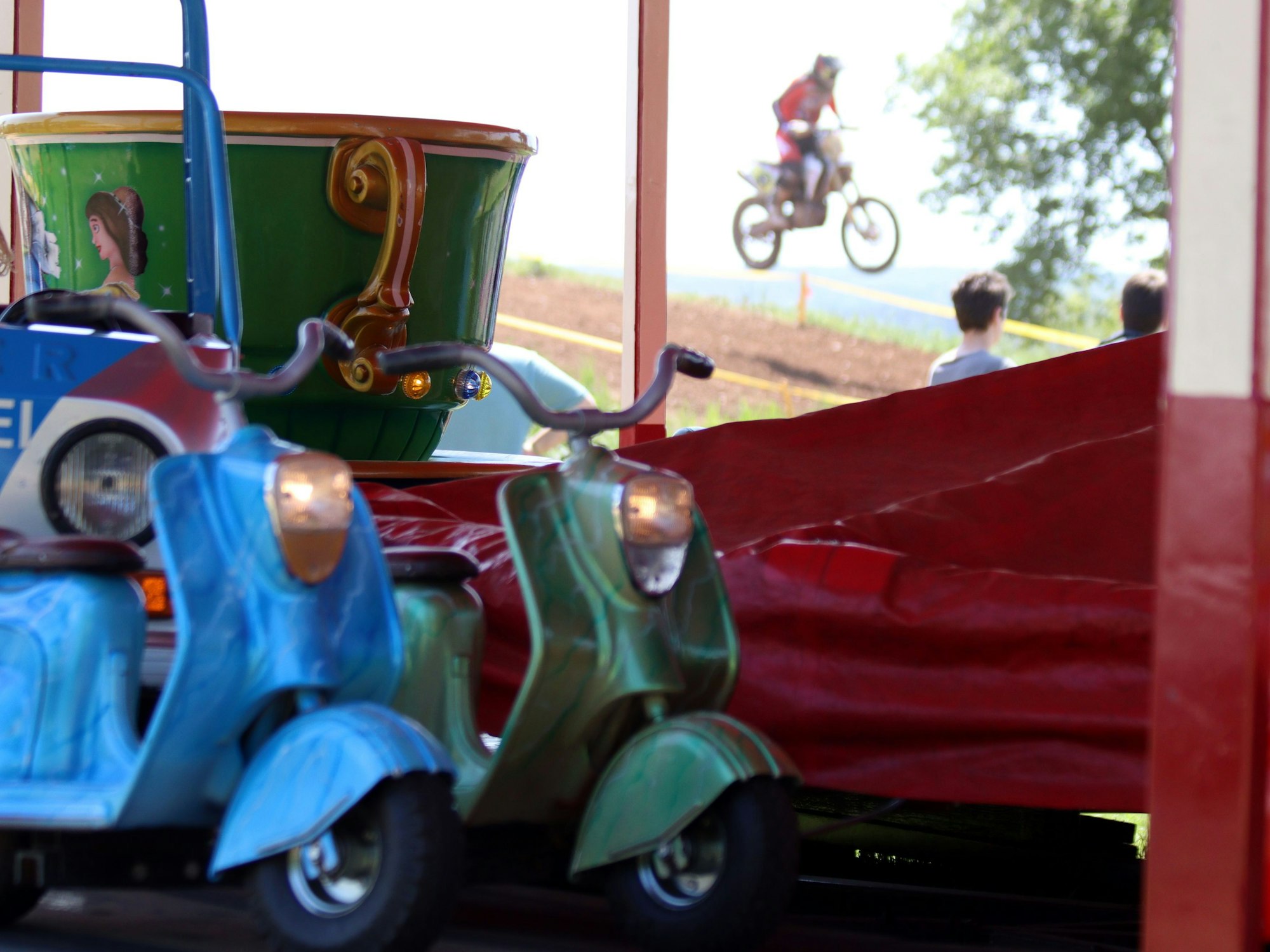 Im Vordergrund Motorroller auf einem Kinderkarussell, im Hintergrund springen die Motocross-Maschinen über die Piste.