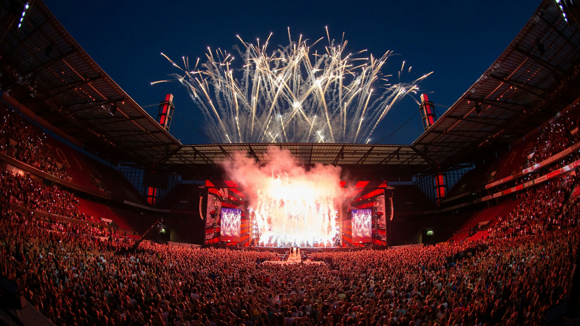 Über dem Rheinenergiestadion explodieren Feuerwerkskörper, die das Publikum im Stadion in rotes Licht tauchen.