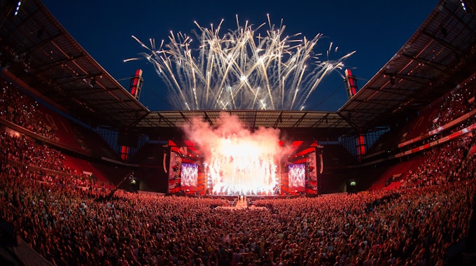 Über dem Rheinenergiestadion explodieren Feuerwerkskörper, die das Publikum im Stadion in rotes Licht tauchen.