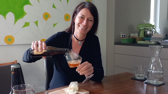 Biersommelière Jutta Knoll aus Bonn-Mehlem schenkt grinsend Bier in ein Glas ein, während sie am Tisch in einer Küche sitzt.
