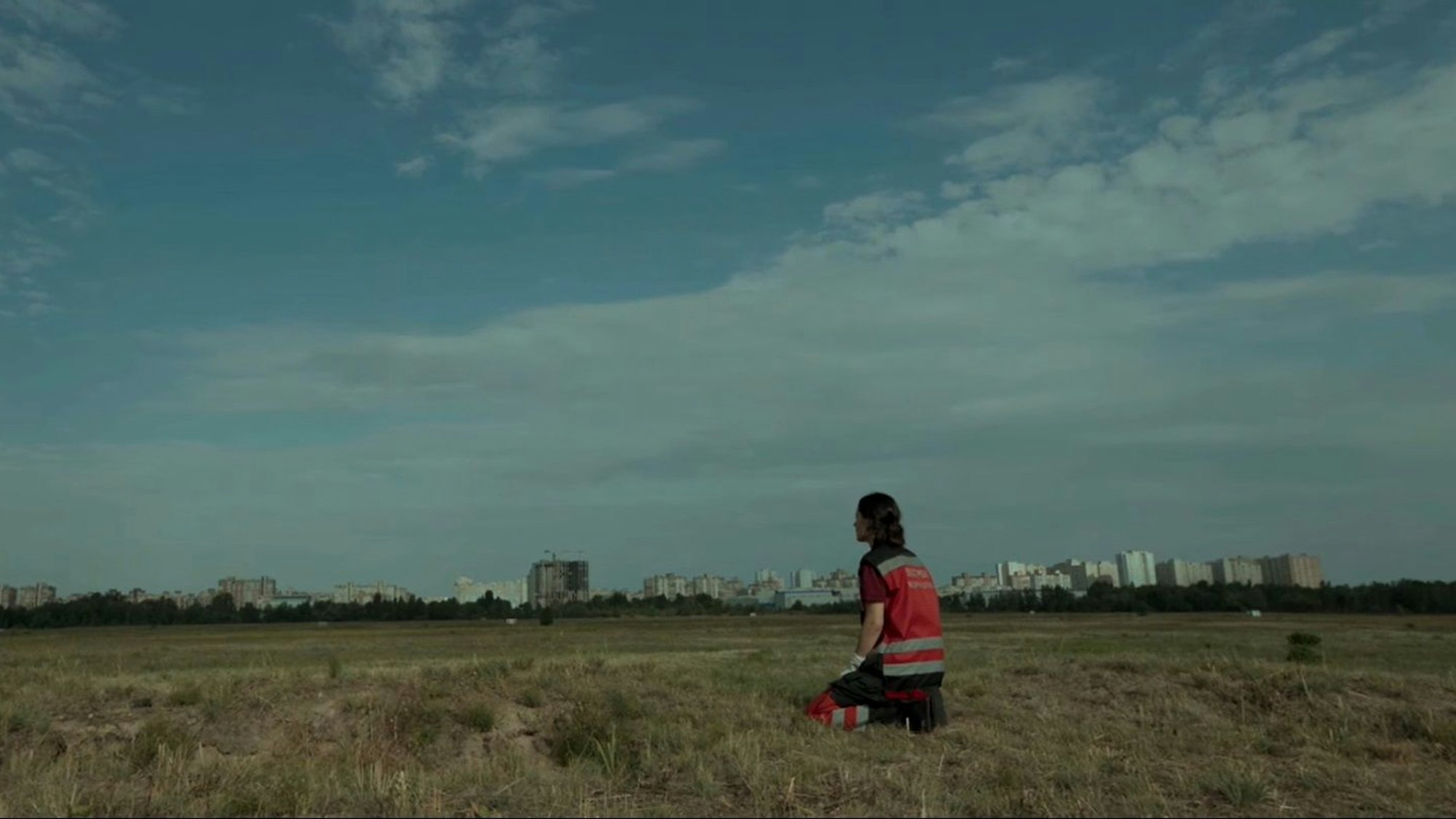 Katia sitzt mit einer roten Warnweste auf einer Wiese, im Hintergrund eine Stadtsilhouette