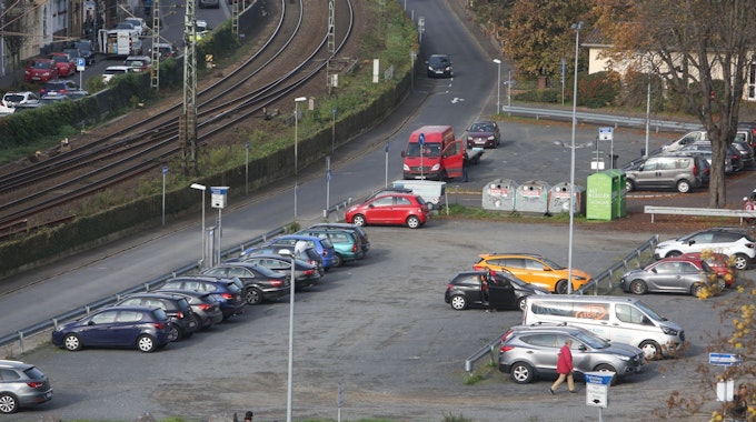 Parkende Autos stehen auf einer Schotterfläche. Links am Bildrand verläuft die rechtsrheinische Bahntrasse.