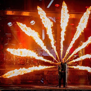 Rammstein-Sänger Till Lindemann steht bei einem Konzert auf der Bühne, er hat die Arme ausgebreitet und singt ins Mikrofon. Auf dem Rücken trägt er einen Pyrotechnik-Apparat, der Flammen zu allen Seiten schießen lässt.