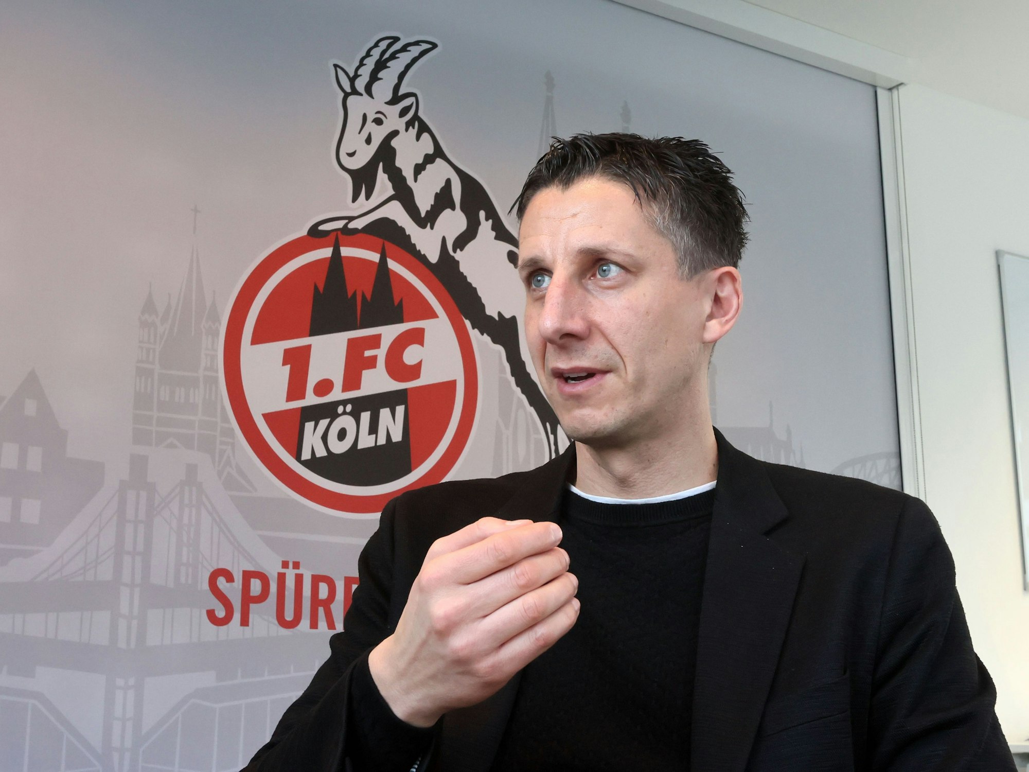 Christian Keller spricht in einem Interview. Hinter ihm das Wappen des 1. FC Köln.