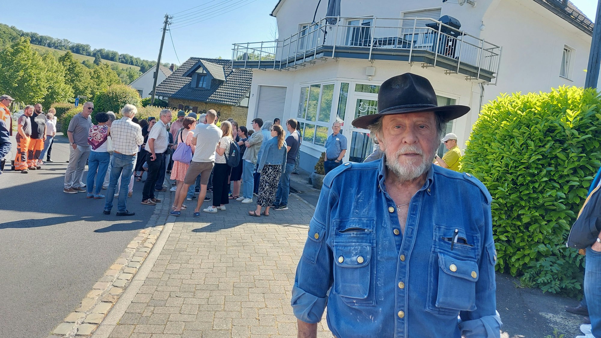 Ein Mann mit Hut und blauem Hemd – der Künstler Gunter Demnig – schaut in die Kamera, im Hintergrund eine Gruppe von Menschen, die an der Stolpersteinverlegung teilnahmen.