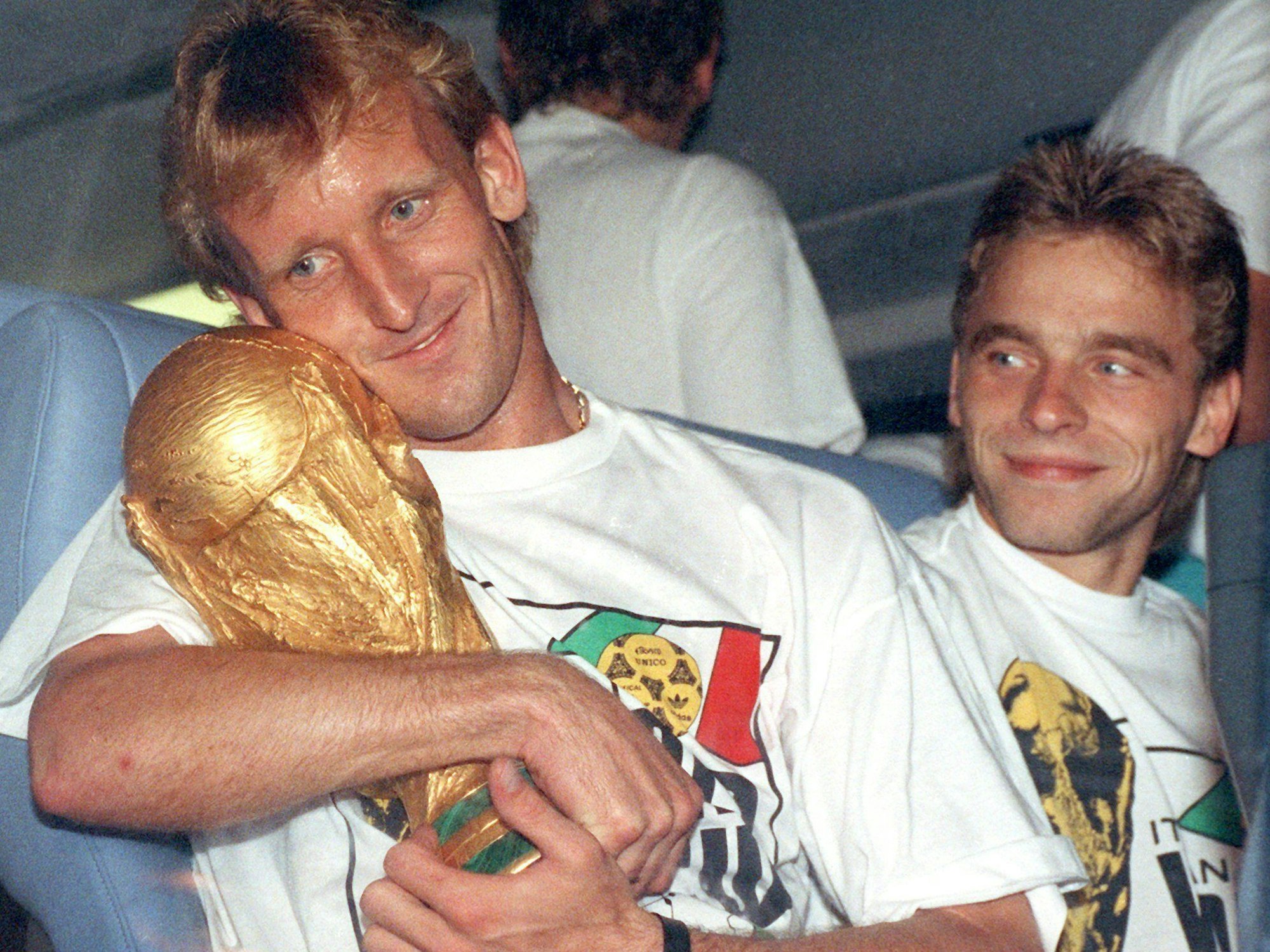 Fußballprofi Thomas Häßler schaut auf dem Rückflug von Rom lächelnd auf seinen Teamgefährten Andreas Brehme, der den WM-Pokal umarmt.