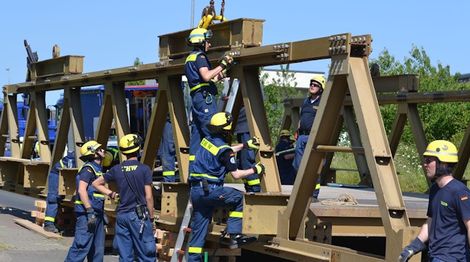 Acht THW-Helfer in blauer Arbeitskleidung und mit gelben Helmen arbeiten am Abbau einer massiven Stahlbrücke.