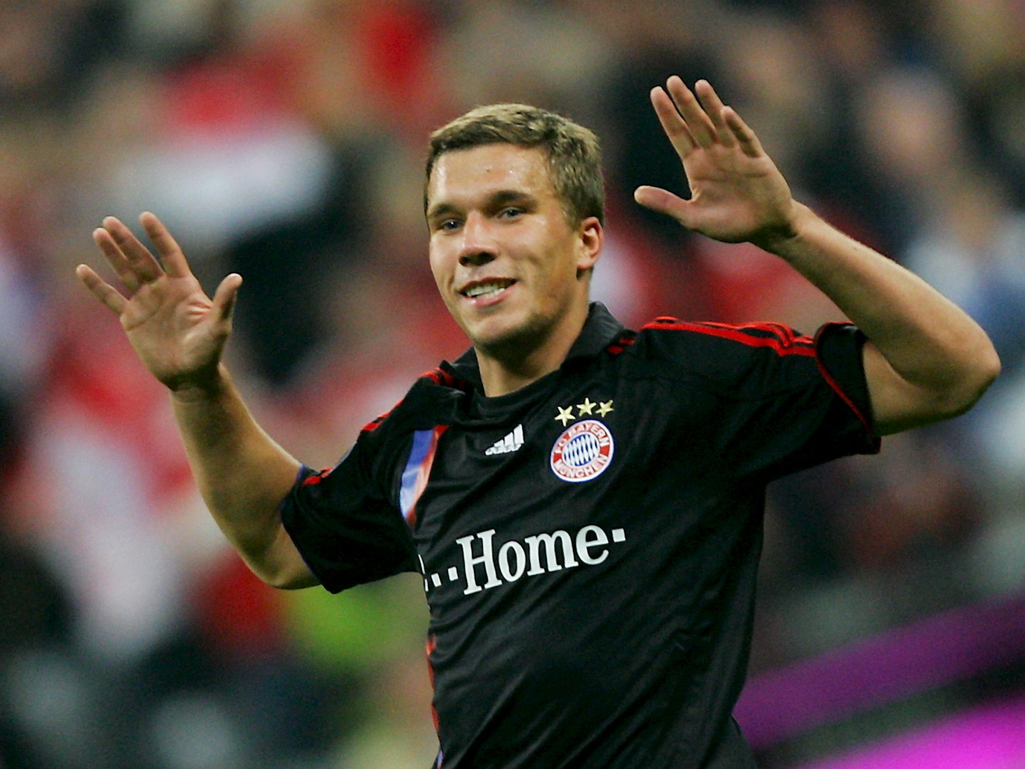 Lukas Podolski trägt ein schwarzes Trikot des FC Bayern München. Er lächelt, seine Hände hebt er in die Höhe.