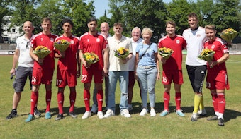 Fußballspieler stehen mit anderen Personen auf einem Fußballfeld und halten Blumen in den Händen.